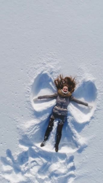 Młoda wesoła kobieta leży na śniegu i robi śnieżne anioły. Zwolnij trochę. Widok z lotu ptaka. Pionowe wideo - Materiał filmowy, wideo