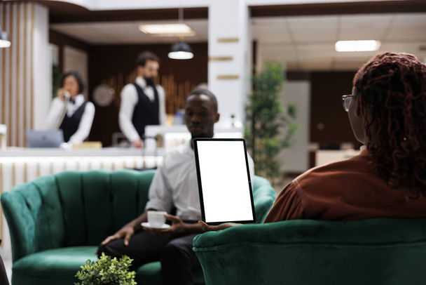 Afroamerikaner im Hotel mit Tablet mit weißem Bildschirm, Blick auf leere Vorlagenanzeige und im Loungebereich sitzend. Weibliche Gast-Haltevorrichtung mit isoliertem Kopierraum. - Foto, Bild