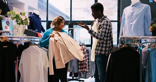 Afrykańska para amerykańska sprawdza stylowy towar, kupuje ubrania w nowoczesnym butiku. Modni klienci analizujący modne stroje w sklepie odzieżowym. Koncepcja mody - Zdjęcie, obraz