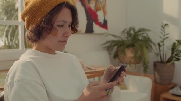 Mediano primer plano de la joven mujer caucásica rizada en gorro sombrero y camiseta blanca sentado en casa, charlando con amigos a través de Internet, escribiendo en el teléfono inteligente y riendo - Imágenes, Vídeo
