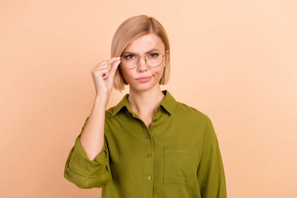 ボブヘアスタイルの不満の不審な女性の写真 緑のシャツの指はベージュ色の背景で隔離された眼鏡を保持します. - 写真・画像