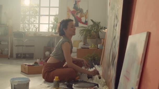 Full portrait shot of happy Caucasian female artist in jumpsuit sitting on floor in workshop and creating large abstract painting, then turning towards camera and smiling - Footage, Video