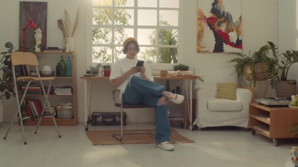 Full shot of young curly Caucasian woman in beanie hat, white t-shirt, ripped jeans sitting on chair at home or in art studio, typing messages on smartphone, while relaxing after work - Footage, Video