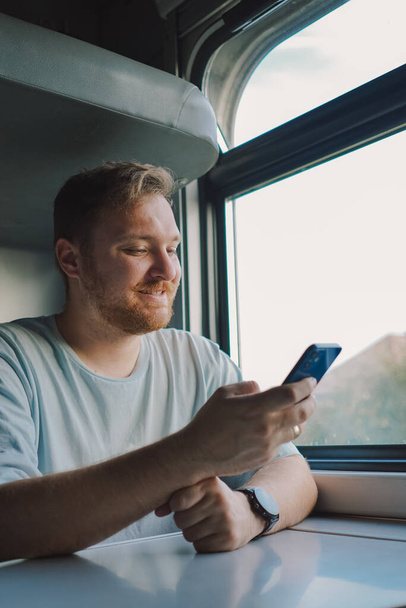 Muž s vousy a knírem v modrém tričku používá smartphone, když cestuje vlakem, sedí ve vlaku a dívá se z okna. - Fotografie, Obrázek