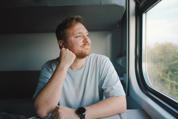 Een man met een baard en snor in een blauw t-shirt op reis per trein, zittend in de trein en kijkend uit het raam. - Foto, afbeelding
