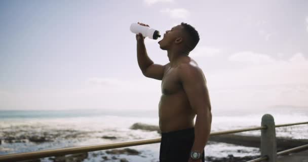 Descanso, fitness y un hombre negro bebiendo agua en la playa después de correr, cardio o ejercicio. Salud, sed y un corredor africano o atleta con una botella para beber después de entrenar junto al océano. - Metraje, vídeo