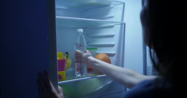 Uma mulher abre um frigorífico à noite e toma uma garrafa de água. Água potável - Filmagem, Vídeo