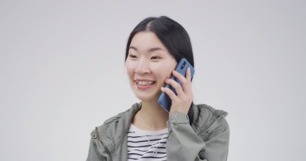 Mujer asiática, sonrisa y llamada telefónica en el estudio para la comunicación, el contacto de audio y noticias sobre fondo blanco. Modelo feliz, teléfono inteligente y chat móvil para la conexión, redes sociales o hola a los chismes. - Metraje, vídeo