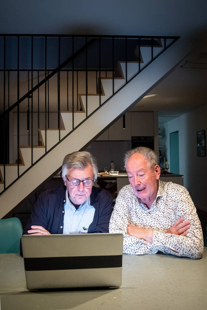 Wciągający obraz pokazujący dwóch starszych mężczyzn, być może przez całe życie przyjaciół, skupionych na ekranie laptopa, uczących się lub dyskutujących razem nad treścią. Nowoczesne wnętrze ze stylowymi schodami w - Zdjęcie, obraz