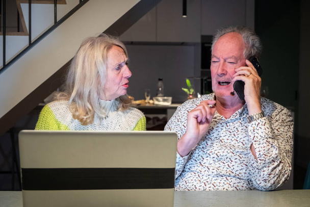 Dieses Bild zeigt ein älteres Paar, das zusammenarbeitet, um ein Problem zu lösen, wobei die Frau besorgt oder verwirrt wirkt, während der Mann am Telefon ist und möglicherweise mit dem Kundendienst spricht. Der Mann - Foto, Bild