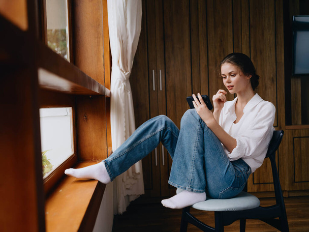 Młoda kobieta siedzi przy oknie z telefonem w ręku uśmiech patrząc online, wiosenny nastrój, dom proste ubrania, domowa przytulna atmosfera, estetyczny styl życia. Wysokiej jakości zdjęcie - Zdjęcie, obraz