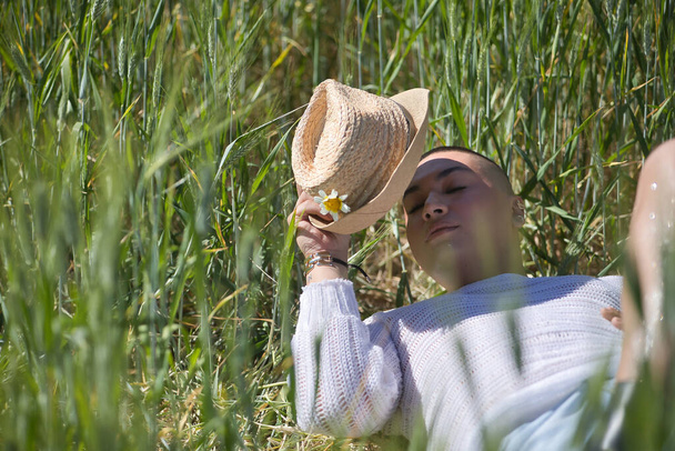 Nicht binäre Person, jung und südamerikanisch, sehr geschminkt, liegt inmitten eines grünen Weizenfeldes, bedeckt sich mit einem Hut vor der Sonne, die Augen geschlossen. Konzeptkönigin, lgbtq +, Stolz. - Foto, Bild