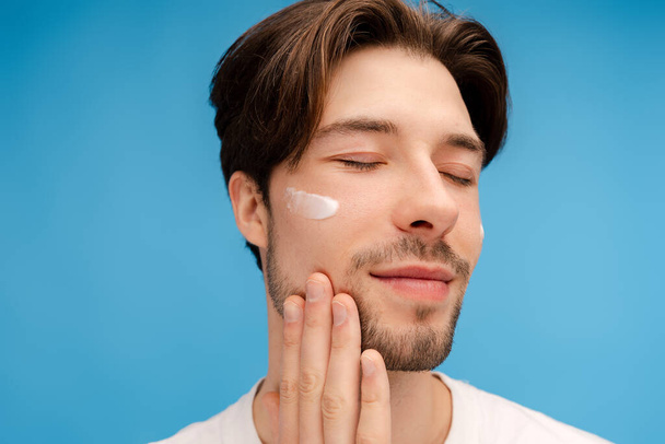 Close up of smiling young man applying lotion to his face. Morning routine. Isolated on blue background. Skin care, cosmetic, spa treatment concept - Fotó, kép
