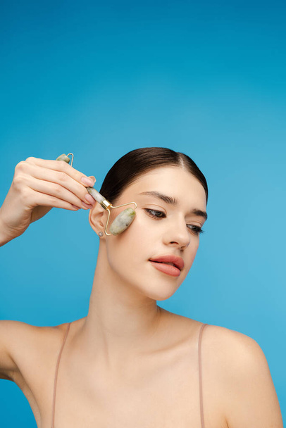 Portrait of young woman in studio using spa jade stone tools for healthy glow, massaging cheekbone. Isolated on blue background. Skin care, anti aging procedure - Photo, Image