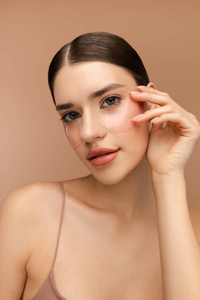 Close up of attractive young woman doing anti-aging skincare routine while using hydrogel eye patches. Isolated on beige background. Skin care, natural beauty, spa treatment concept - Photo, Image