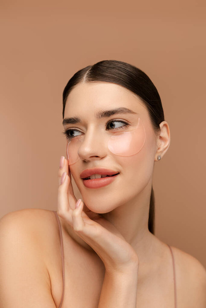 Portrait of smiling young lady posing at camera while using hydrogel eye patches. Isolated on beige background. Skin care, natural beauty, spa treatment concept - Photo, Image