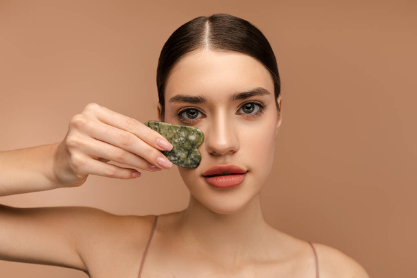 Portrait of pretty young lady using gua sha stone scraper while doing skin care routine. Isolated on beige background. Anti-Aging skincare routine - Fotoğraf, Görsel