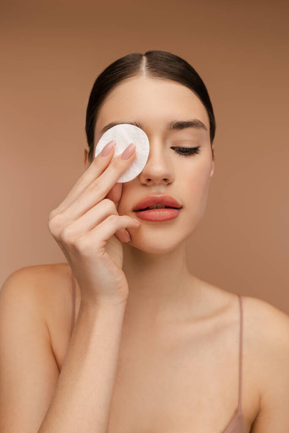 Portrait of beautiful lady removing make-up and cleaning her skin, holding the sponge in front of her eye, posing in studio. Isolated on beige background. Perfect cosmetology skin care - Photo, Image