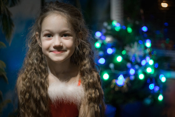 Een klein meisje met een mooie glimlach. Blond harig kind viert Kerstmis met feestelijke decoraties en boom. Zachte selectieve focus.  - Foto, afbeelding