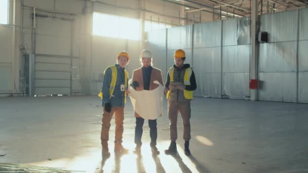 Ampia ripresa di uomo d'affari caucasico in abiti casual holding piano di costruzione e discutere la sua attuazione con due lavoratori edili maschili Biracial nel centro di un nuovo edificio spazioso - Filmati, video