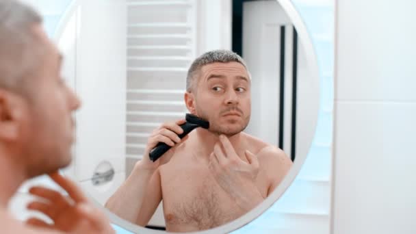 Вид сзади мужского лица в зеркале в ванной обнаженного парня с каблуком, мужчина брюнет использует электрическую бритву для бритья волос от подбородка легкого бритья дома - Кадры, видео