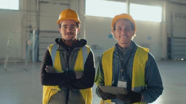 Середній портрет двох чоловіків Бірао будівельників в твердих капелюхах і захисний робочий одяг посміхається і позує на камеру з планшетом в руках під час робочого дня всередині порожньої просторої будівлі - Кадри, відео