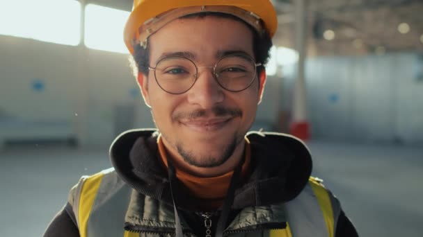 Szoros portré boldog Biracial szakállas építőmunkás szemüvegben és biztonsági kalap nézi kamera munkanapon belül egy nagy üres épületben a napsütéses napon - Felvétel, videó