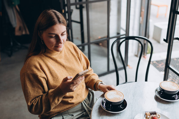 Gyönyörű fiatal nő alkalmi ruhában mosolyog, iszik egy csésze kávét és egy telefont tart a kezében, miközben a kávézóban tölti az idejét. - Fotó, kép
