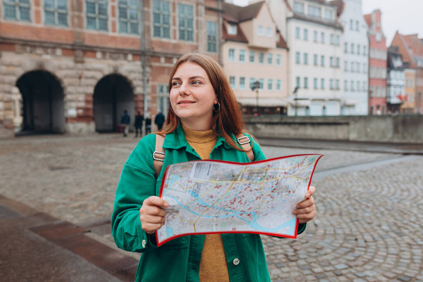 Attraktive junge Touristinnen erkunden die neue Stadt. Rotschopfiges Mädchen mit Rucksack, das eine Landkarte aus Papier in der Danziger Straße hält. Europa im Herbst bereisen - Foto, Bild