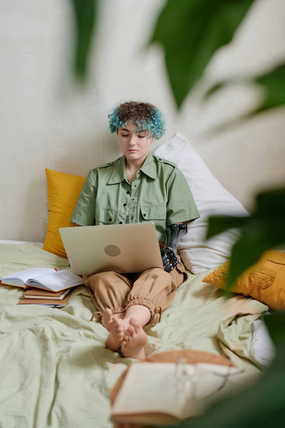 Έξυπνο κορίτσι με προσθετικό χέρι κάθεται στο κρεβάτι και εργάζεται σε φορητό υπολογιστή - Φωτογραφία, εικόνα