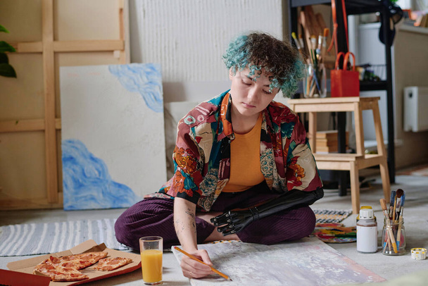 Giovane donna creativa con braccio protesico che mangia pizza quando disegna su tela - Foto, immagini
