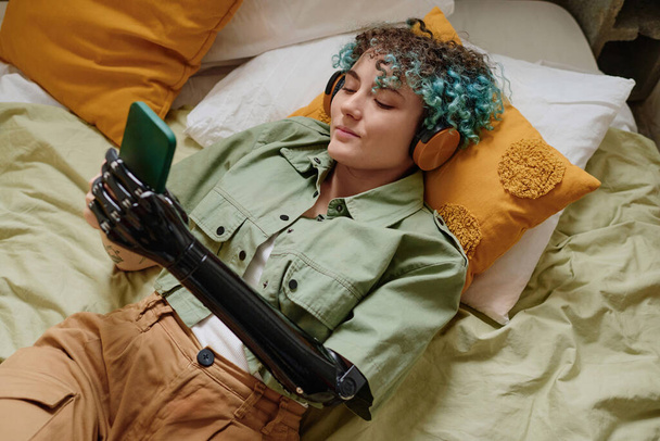 Mujer joven sonriente con brazo protésico escuchando música en los auriculares y revisando las redes sociales en el teléfono inteligente - Foto, imagen