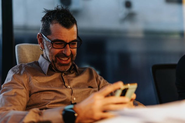 Pohledný muž středního věku, který po pracovní době kontroluje telefon a naslouchá rozhovoru svých kolegů a usmívá se. - Fotografie, Obrázek