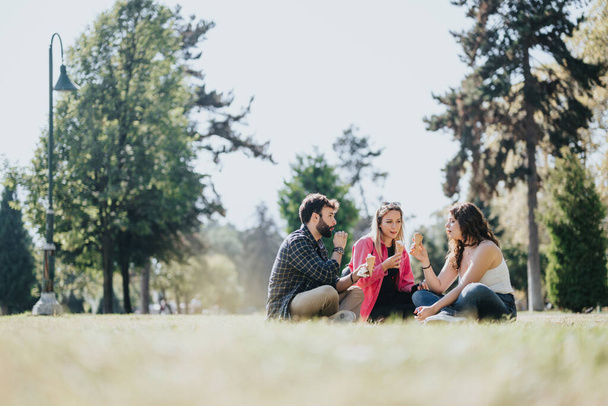 Mladí přátelé si užívají bezstarostný slunečný den v městském parku. Smějí se, mluví a odpočívají na trávě, vychutnávají si zmrzlinu a pozitivní energii svého volného času. - Fotografie, Obrázek