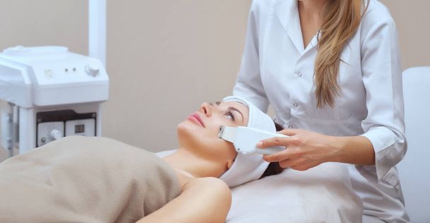 Lékař-kosmetolog provádí ultrazvukové čištění pleti krásné mladé ženy v salonu krásy. Kosmetologie a profesionální péče o pleť. - Fotografie, Obrázek