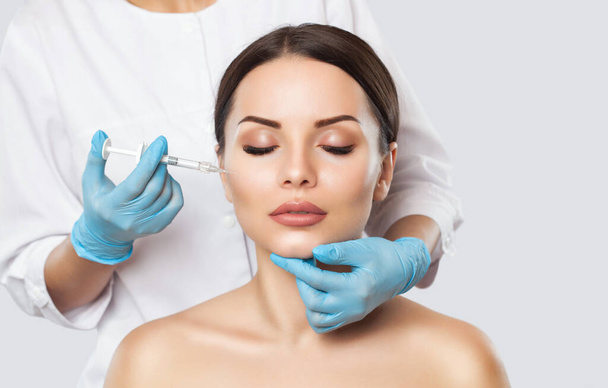Doktor kozmetik uzmanı güzellik salonundaki genç ve güzel bir kadının cildindeki kırışıklıkları sıkmak ve düzleştirmek için yüz enjeksiyonlarını yenileme prosedürünü uyguluyor.. - Fotoğraf, Görsel