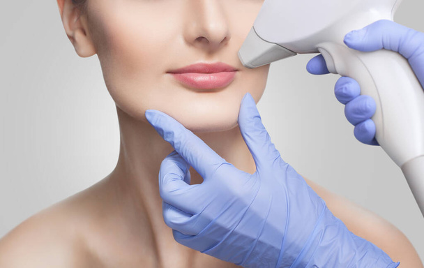 De cosmetoloog doet de procedure voor laser ontharing van ongewenste haren van het gezicht aan een jong meisje in een schoonheidssalon. - Foto, afbeelding