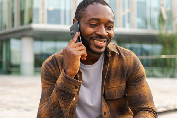 African american man talking on smartphone on street in city outdoor. Man with cell phone chatting with friends. Smiling person making answering call by cellphone. Guy having conversation by mobile - Photo, Image