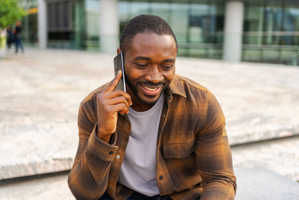 Африканський американець розмовляє по смартфонах на вулиці в місті на відкритому повітрі. Чоловік з мобільним телефоном спілкується з друзями. Усміхнена людина робить відповідь на дзвінок по мобільному телефону. Хлопець розмовляє по мобільному телефону - Фото, зображення