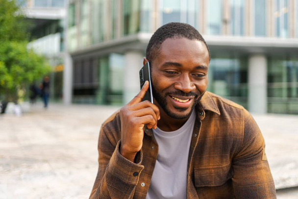 Африканський американець розмовляє по смартфонах на вулиці в місті на відкритому повітрі. Чоловік з мобільним телефоном спілкується з друзями. Усміхнена людина робить відповідь на дзвінок по мобільному телефону. Хлопець розмовляє по мобільному телефону - Фото, зображення