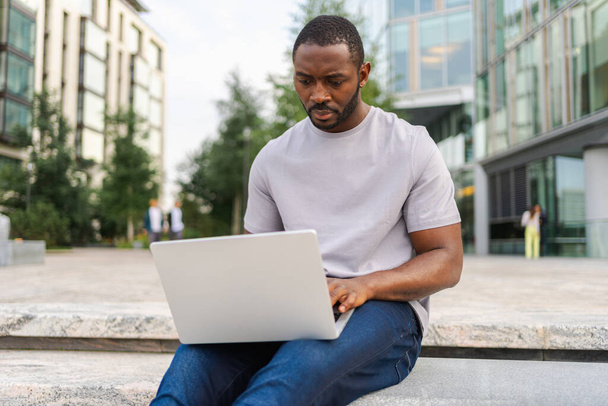 Afroamerykanin freelancer za pomocą laptopa wpisując na czacie na ulicy miejskiej w mieście. Facet ma wirtualne spotkanie na czacie online. Programista przedsiębiorczości pracujący na laptopie na zewnątrz. Praca zdalna - Zdjęcie, obraz