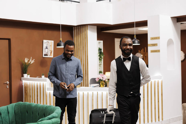 Souriant sympathique groom de l'hôtel aidant les clients avec ses bagages après la procédure d'enregistrement, le gars afro-américain groom tirant valise livrant des bagages dans la chambre. Personnel de bureau Bell - Photo, image