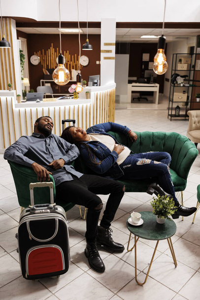Усталая пара, спящая в вестибюле отеля после долгих международных рейсов, страдает от усталости от путешествий и смены часовых поясов. Вымотанные туристы дремлют на диване в ожидании регистрации на курорте - Фото, изображение