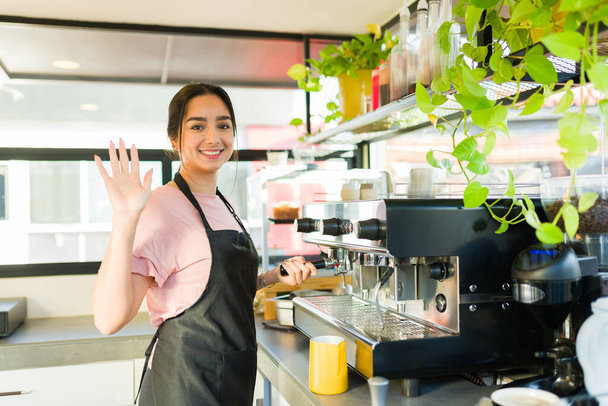 Mujer latina feliz y barista saludando a un cliente mientras sonríe y trabaja preparando café o capuchino - Foto, imagen