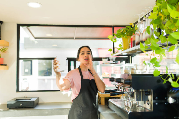 Hermosa mujer latina y barista llamando el nombre de un cliente mientras entrega un café para ir a la cafetería - Foto, Imagen
