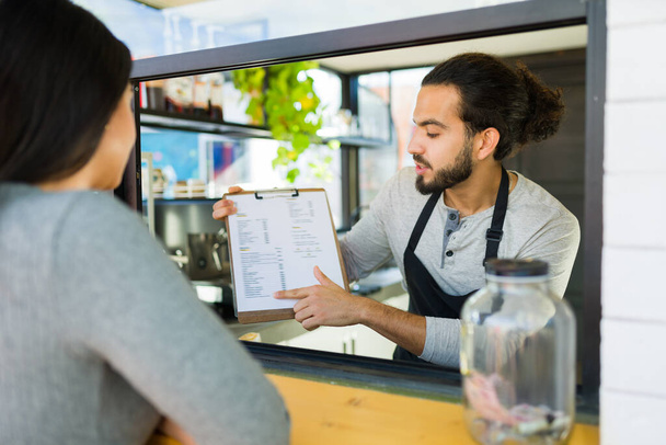 Όμορφος μπάρμαν που δείχνει το μενού του καφέ σε μια πελάτισσα που παραγγέλνει καφέ και φαγητό στην καφετέρια - Φωτογραφία, εικόνα