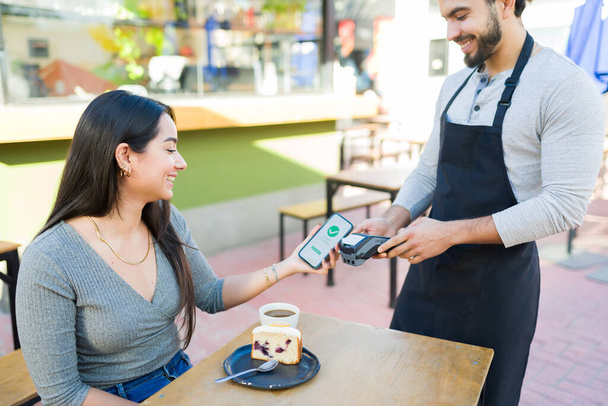 Ενθουσιασμένη νεαρή γυναίκα που πληρώνει για τον καφέ και τη ζαχαροπλαστική της χρησιμοποιώντας την εφαρμογή smartphone της ενώ χαμογελάει στον εργάτη του καφέ - Φωτογραφία, εικόνα