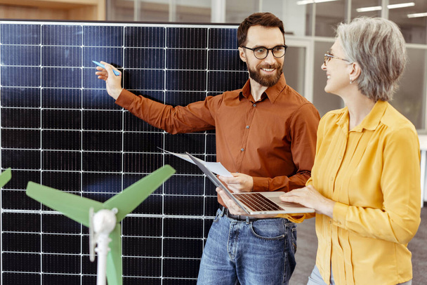 Smiling bearded man and senior woman, engineers, developers talking, holding laptop, planning project, cooperation using solar panel and windmill electric generator working in modern office. Teamwork - Photo, Image