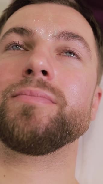 Férfi a maszk kozmetikai eljárás spa szalonban. Felismerhetetlen kozmetikus, aki hidratáló arcpakolást alkalmaz egy fiatal férfi arcán ecsettel. Függőleges videó - Felvétel, videó