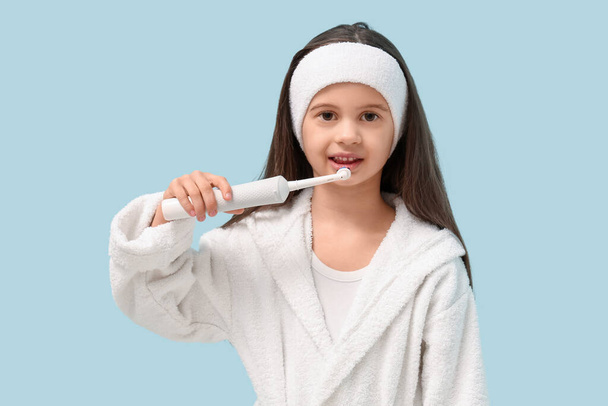 Nettes kleines Mädchen im Bademantel mit elektrischer Zahnbürste auf blauem Hintergrund - Foto, Bild